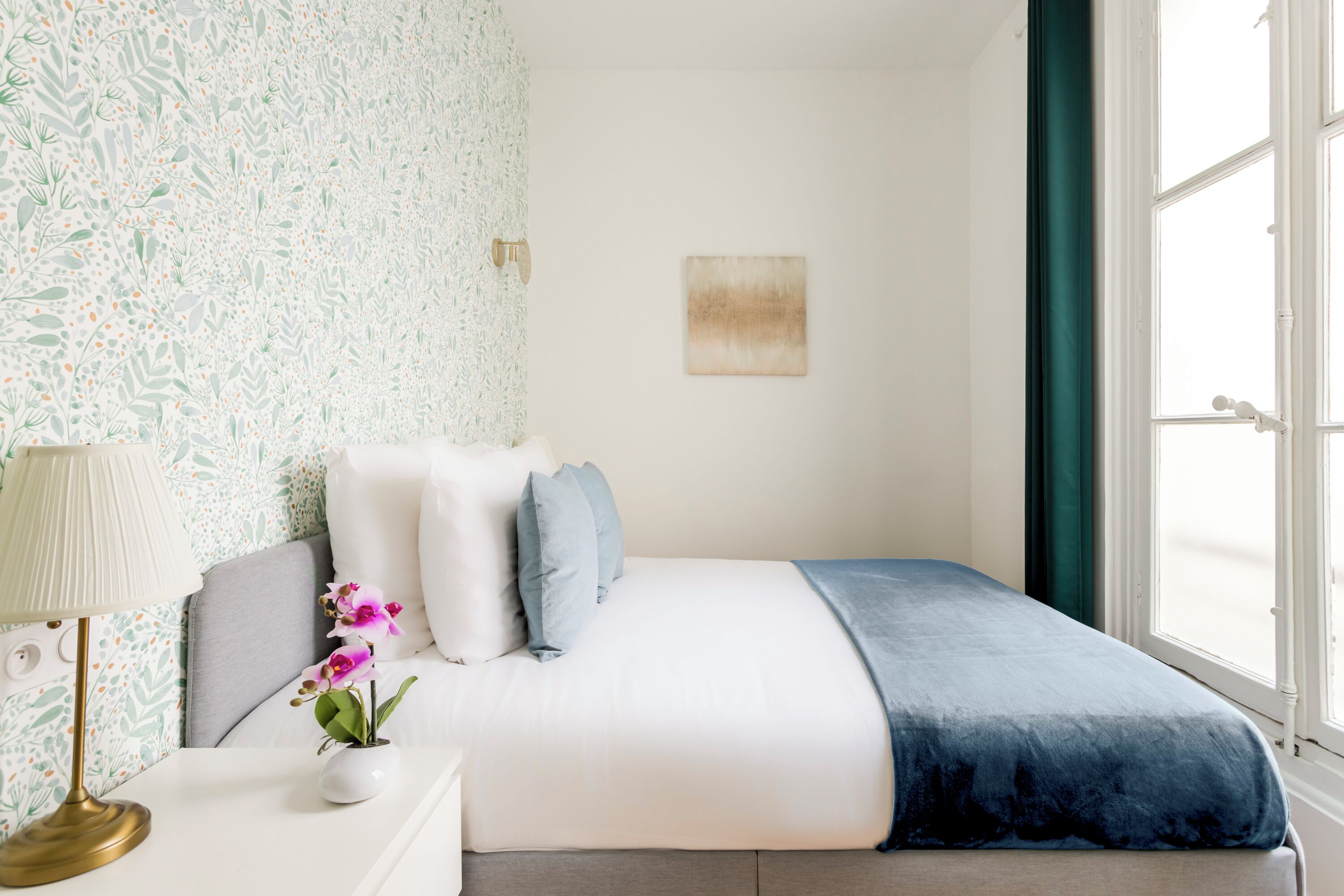 A bedroom with a bed, pillows, a bedside table with a lamp, a vase with flowers, and a window with curtains.