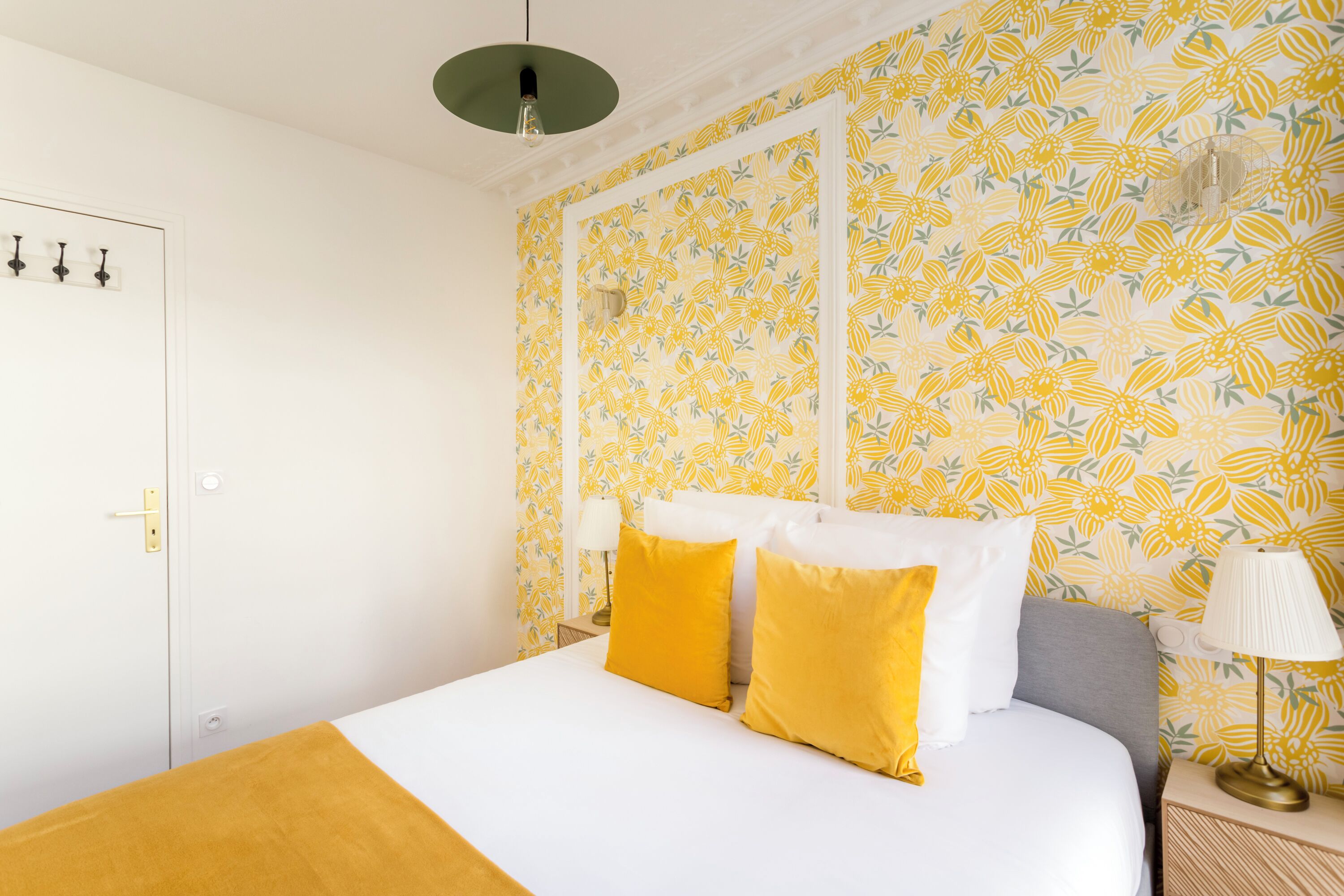 Une chambre d’hôtel avec un lit, des oreillers jaunes, une couvre-lit blanche, une table de chevet avec une lampe et un papier peint à motifs floraux.