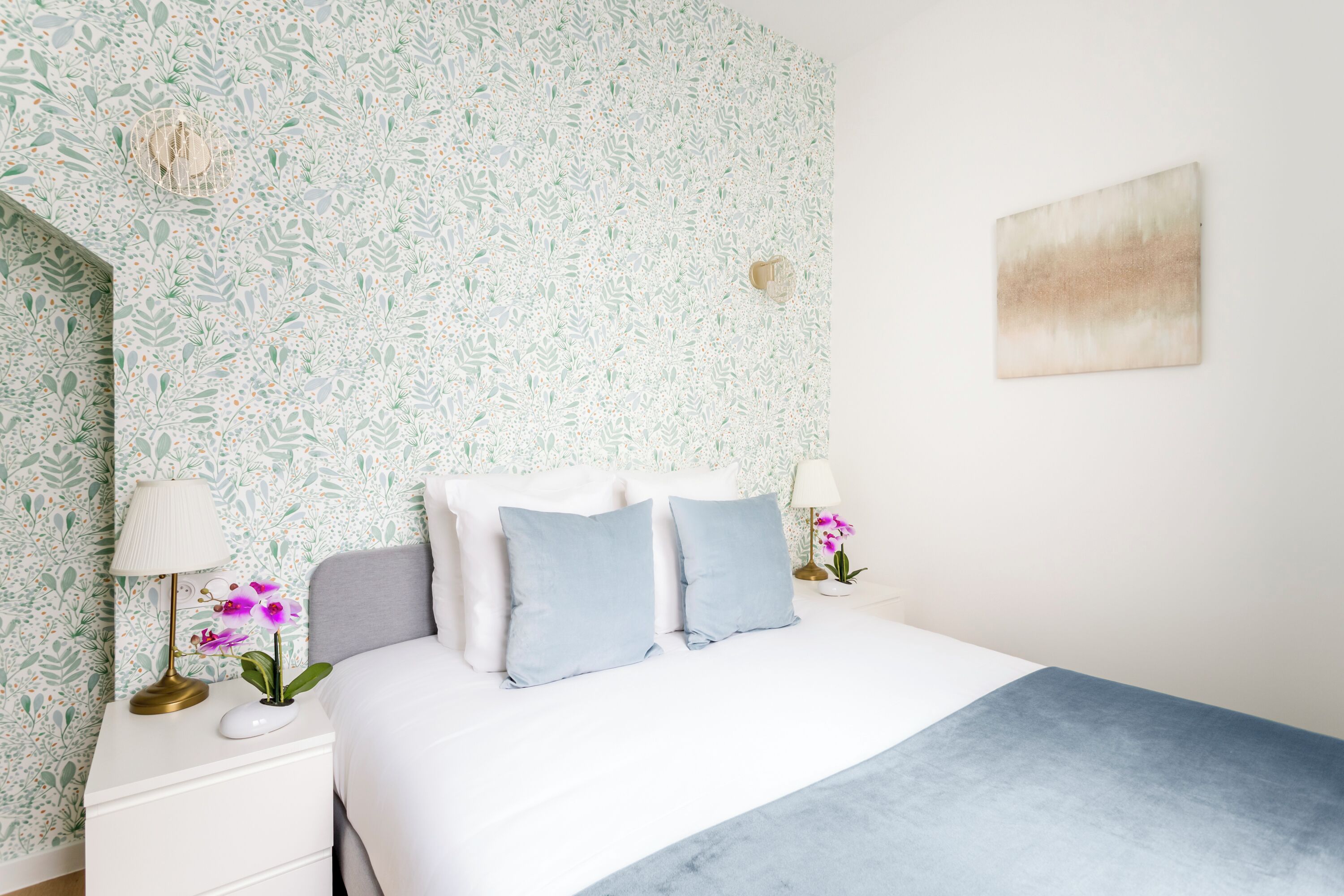 A bedroom with a floral wallpaper, a bed with blue bedding, two bedside tables with lamps, and a single artwork on the wall.