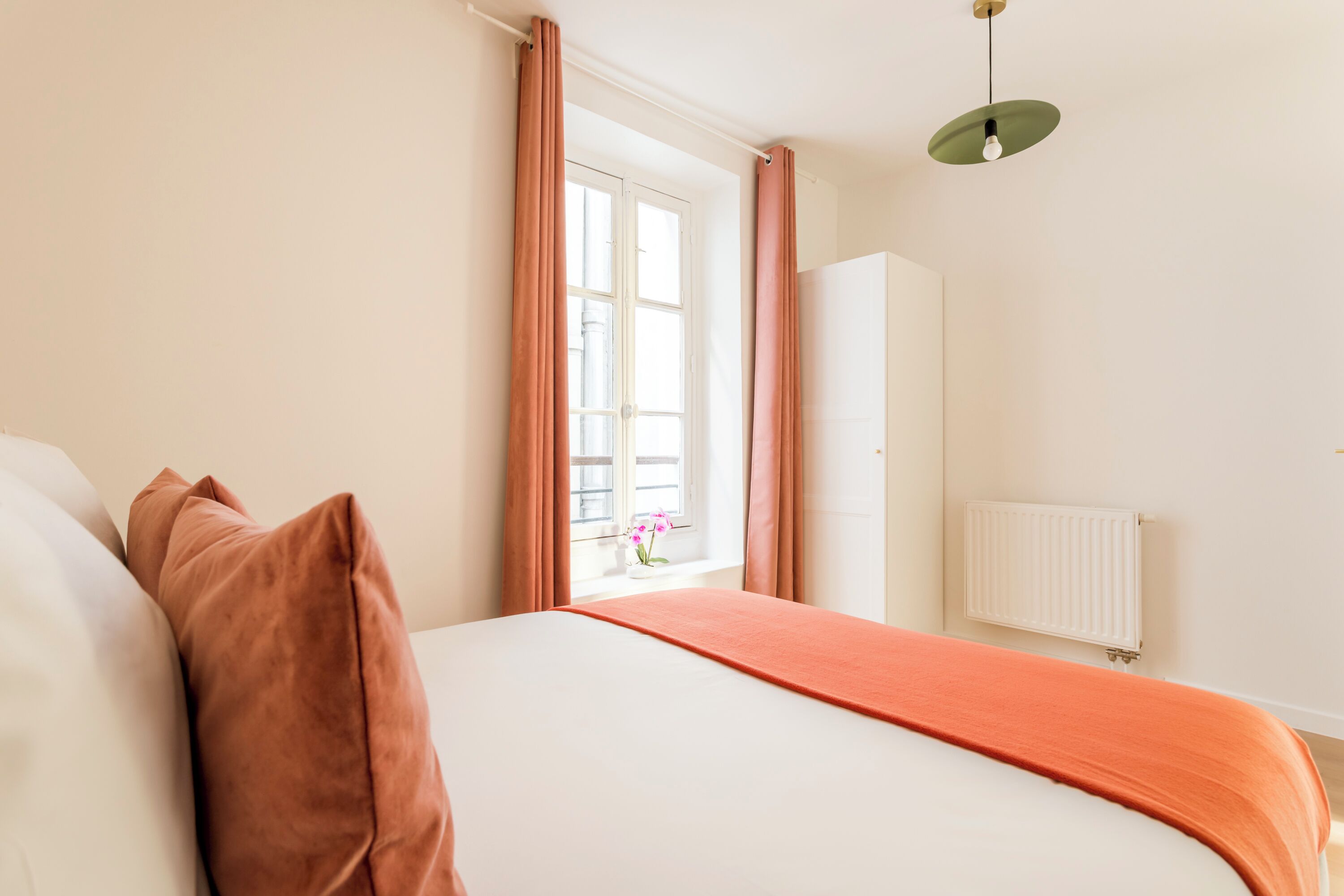 Ein Schlafzimmer mit einem Bett, einem Fenster mit Vorhängen, einem Heizkörper und einer Pendelleuchte.