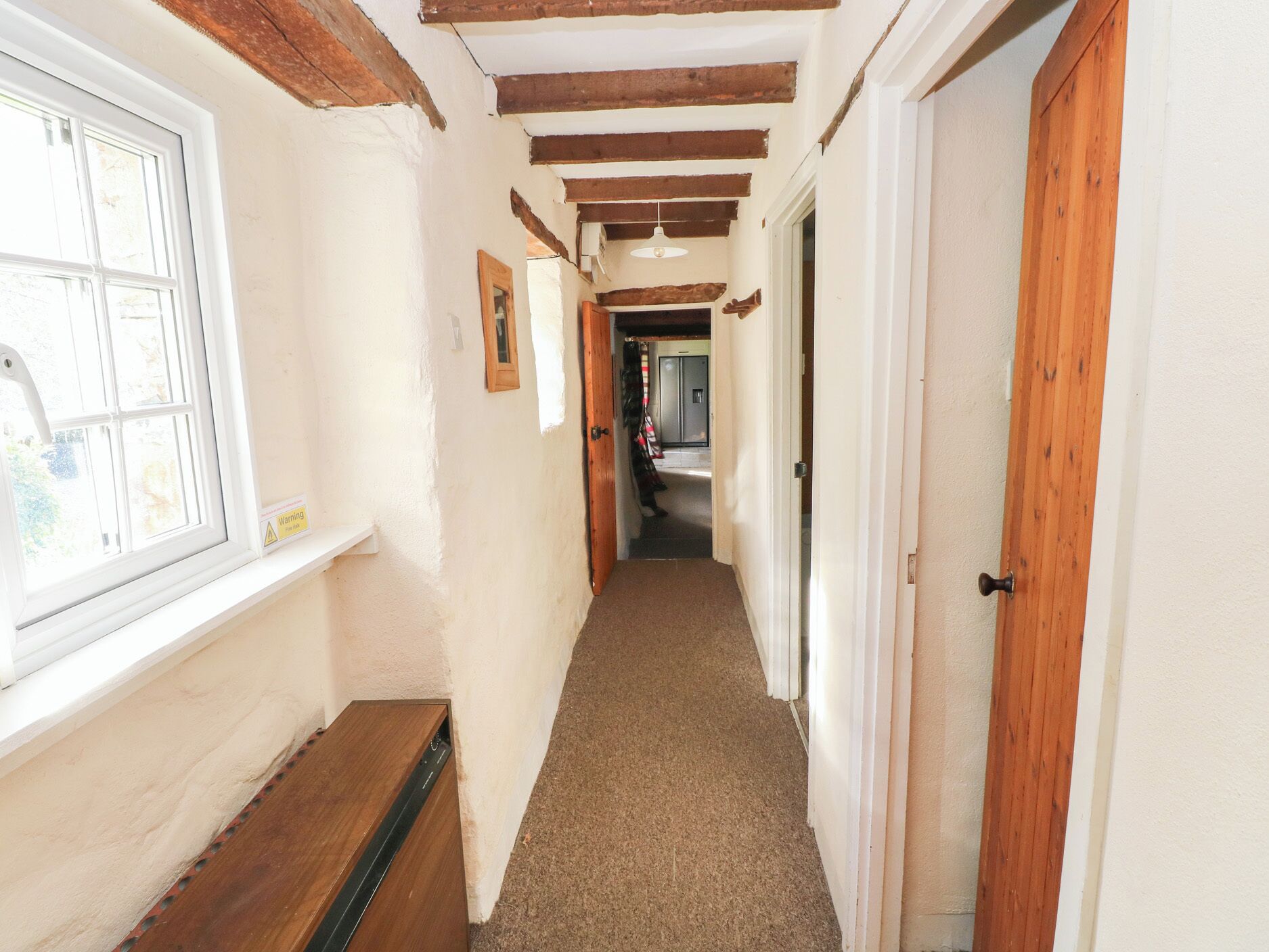 A narrow hallway with wooden beams on the ceiling, a window on the left, and a door on the right.