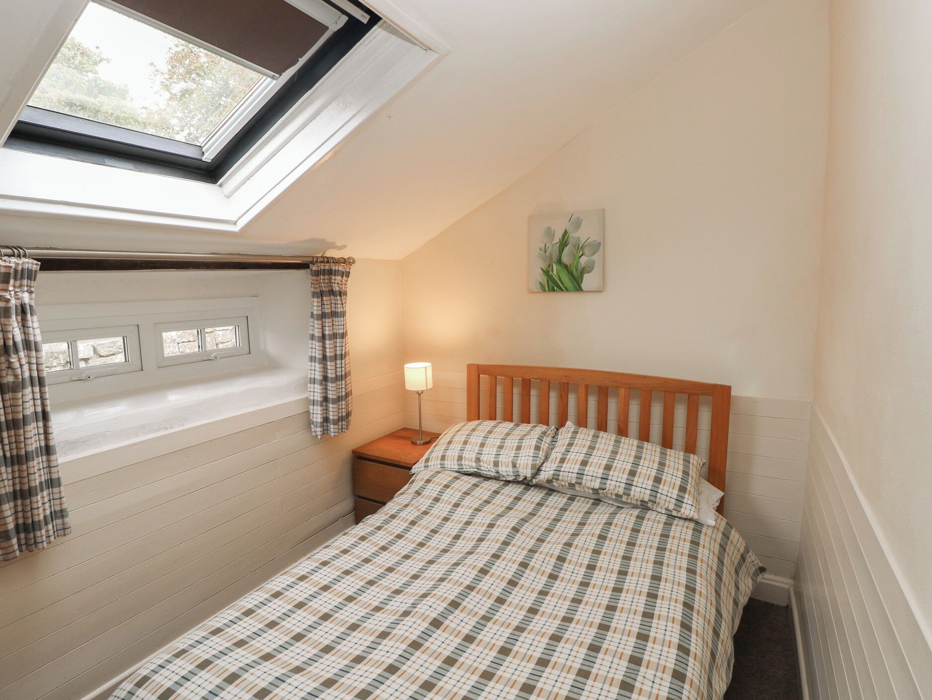 A bedroom with a skylight, a bed with plaid bedding, a nightstand with a lamp, and a painting on the wall.