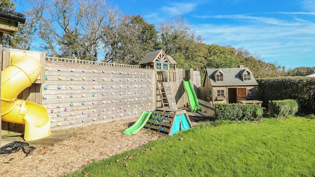 Een achtertuin met een klimmuur, twee glijbanen, een houten speeltuintje en een huis.
