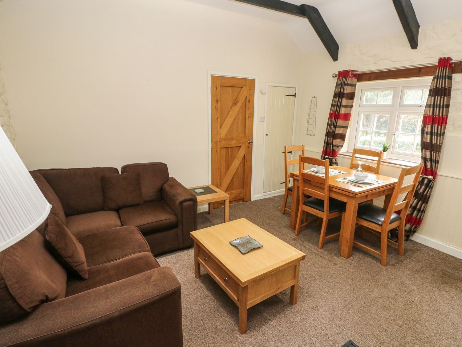 A cozy living room with a brown sofa, a wooden coffee table, and a dining table with chairs. There is a wooden door and a window with curtains.