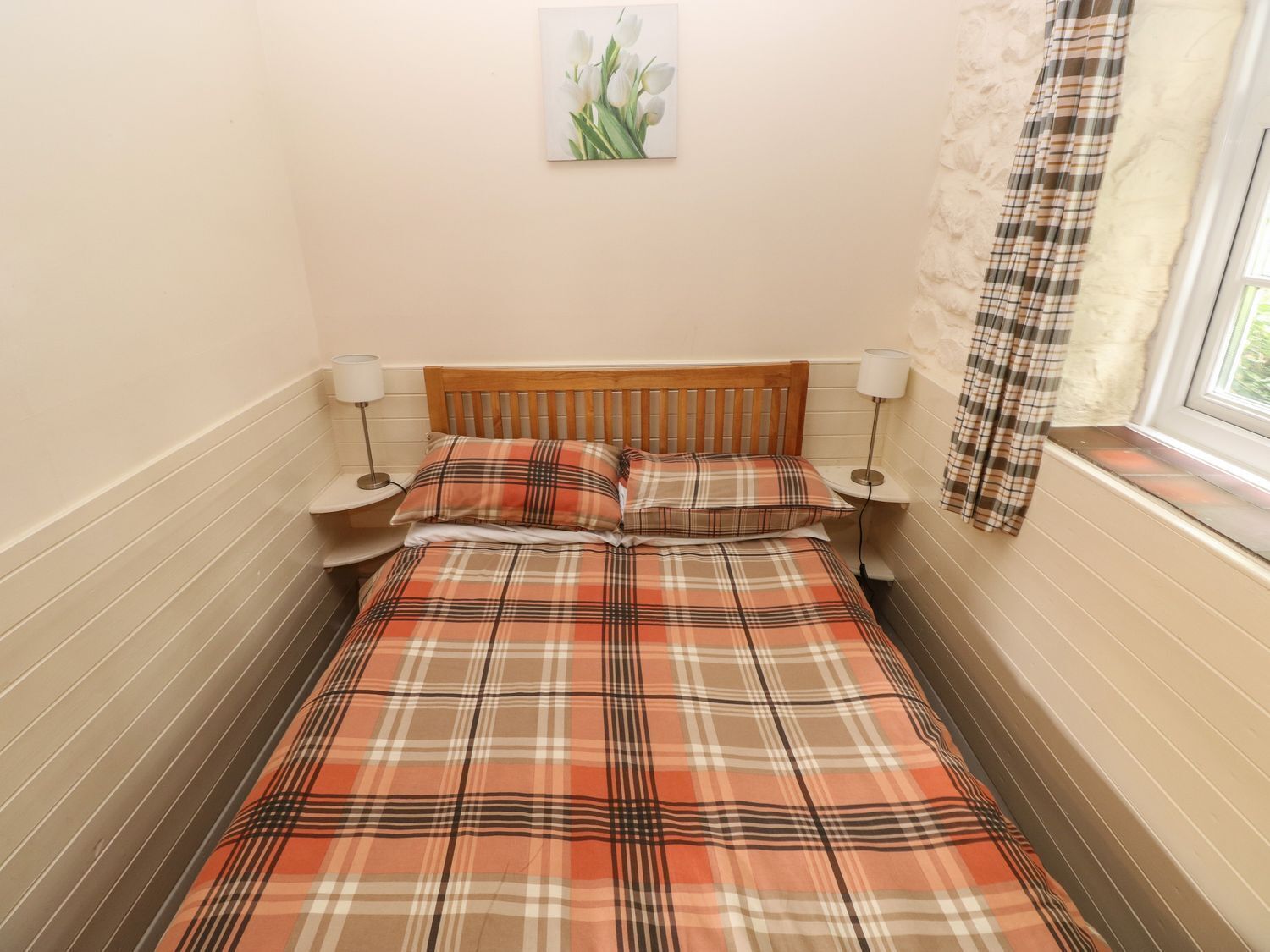 A bed with a plaid bedspread, two bedside lamps, a window with a curtain, and a wall-mounted picture of flowers.