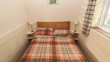 A bed with a plaid bedspread, two bedside lamps, a window with a curtain, and a wall-mounted picture of flowers.