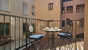 En balkon med et rundt bord, to stole, en lille urtepotte og et vindue med persienner.