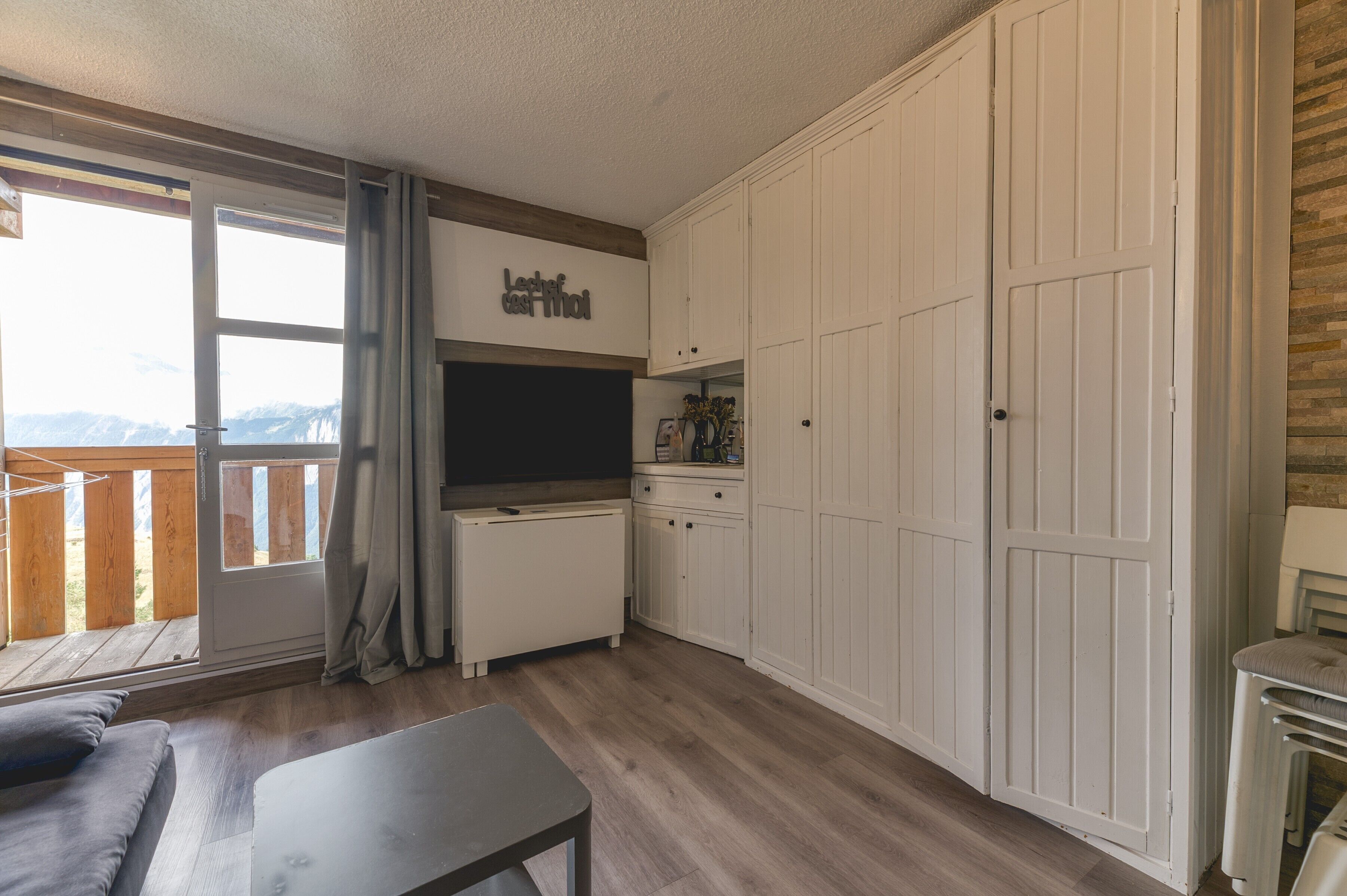 Una habitación con sofá, mesa, zona de cocina con armarios, radiador y puerta corrediza de cristal que da a un balcón exterior.