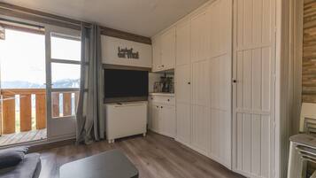 Una habitación con sofá, mesa, zona de cocina con armarios, radiador y puerta corrediza de cristal que da a un balcón exterior.