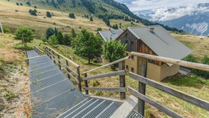 Een houten chalet met een metalen dak, omgeven door een houten hek, met een berglandschap op de achtergrond.