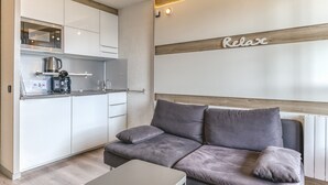 Een moderne kitchenette met een magnetron, koffiezetapparaat en waterkoker. Er staat een grijze bank met kussens en een kleine tafel. Op de muur staat een bordje met de tekst 'Relax'.