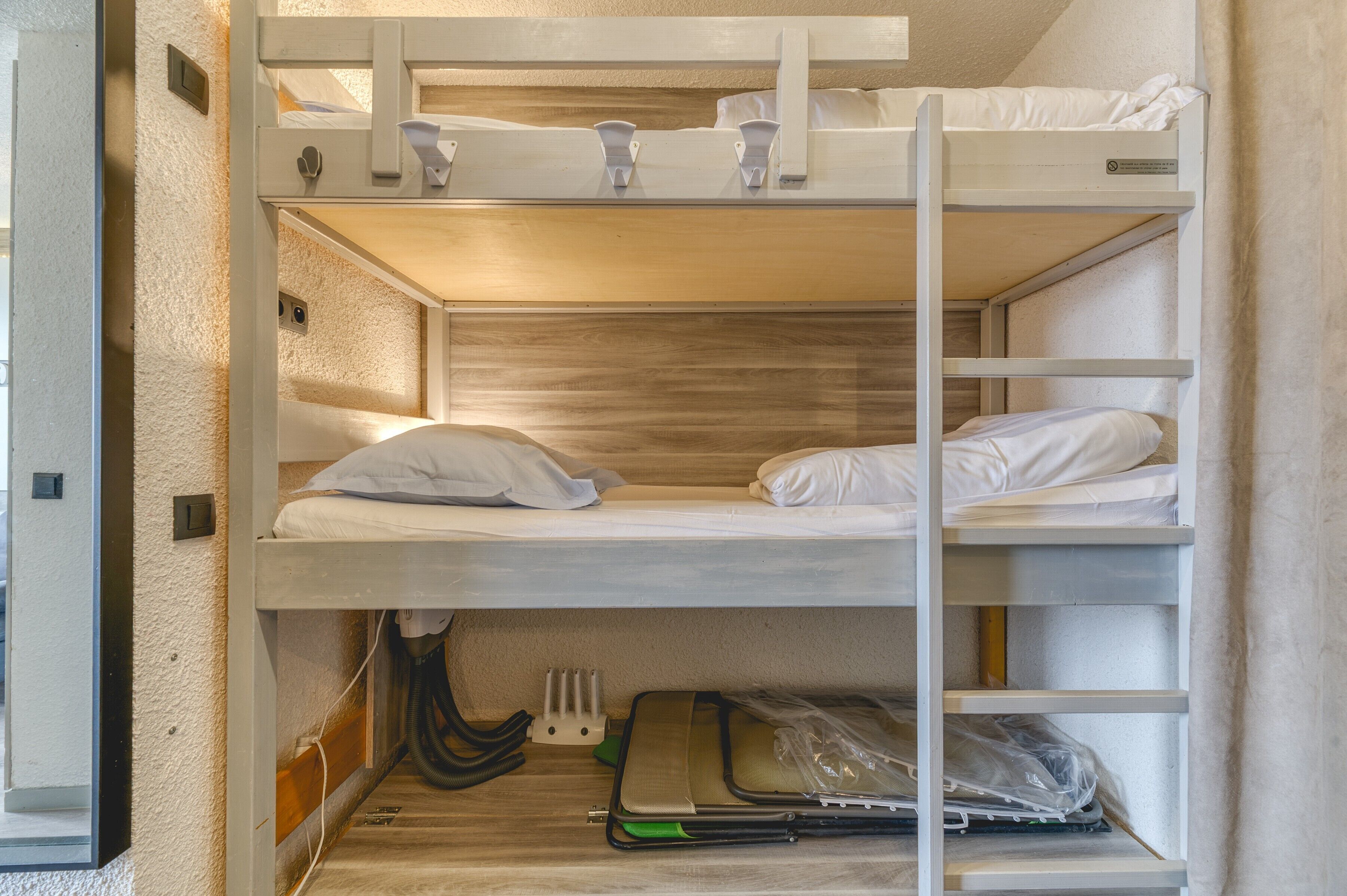 A bunk bed with a wooden headboard, a lower shelf with a laptop and a green notebook, and a power outlet on the wall.