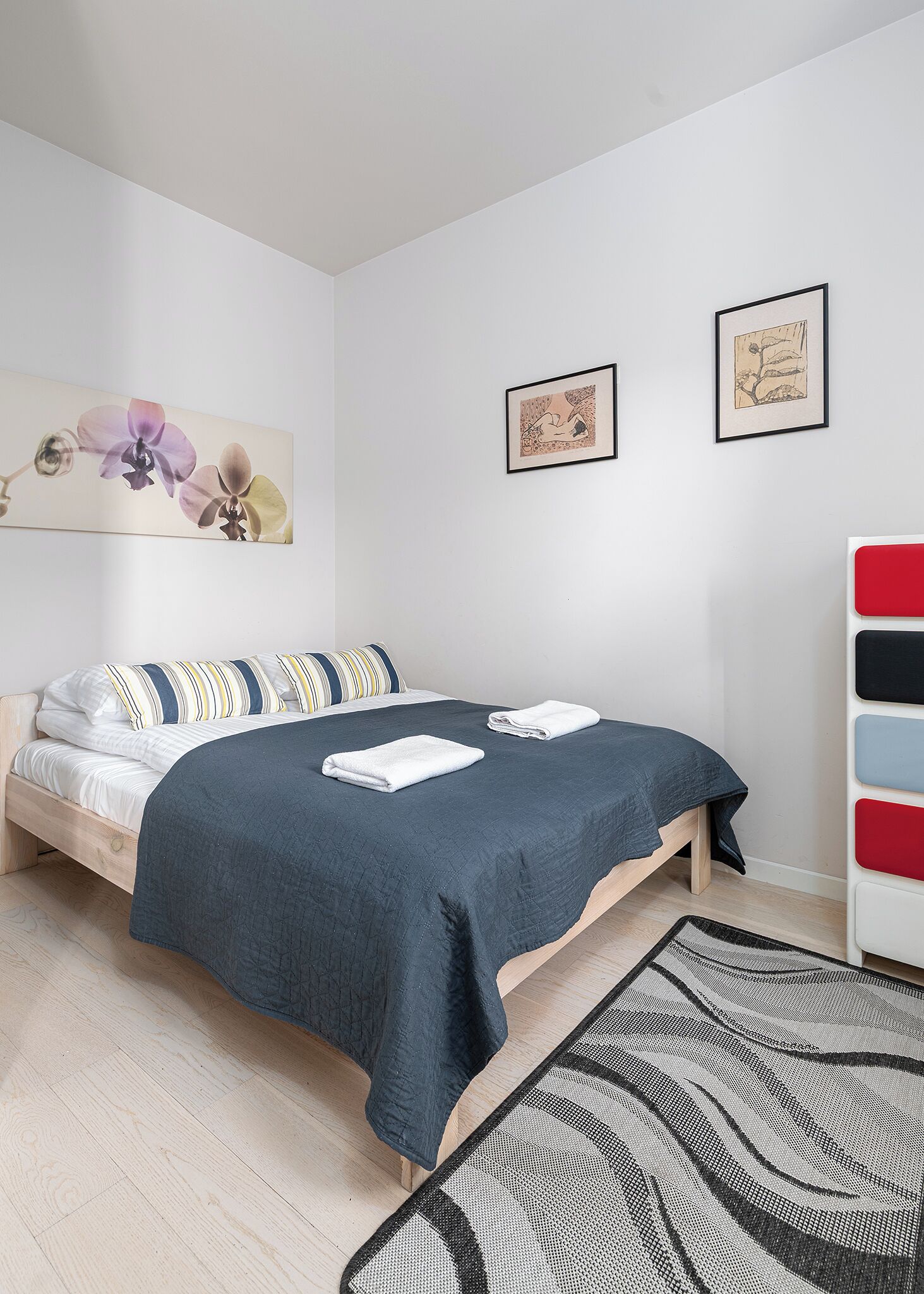 Un lit bien fait, recouvert d’une couvre-lit bleu foncé et agrémenté de deux serviettes blanches pliées, un plancher en bois, un tapis à motifs et deux œuvres d’art encadrées accrochées au mur.