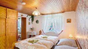 Una habitación con dos camas, techo de madera, papel tapiz floral, una ventana con cortinas de encaje y una mesita con una lámpara.