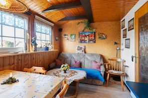 Een gezellige kamer met een houten plafond, een eettafel met een bloemen tafelkleed, een bank met kussens en een kleine houten stoel.