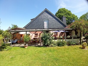Ein Haus mit geneigtem Dach, einer überdachten Terrasse und einem gepflegten Garten.