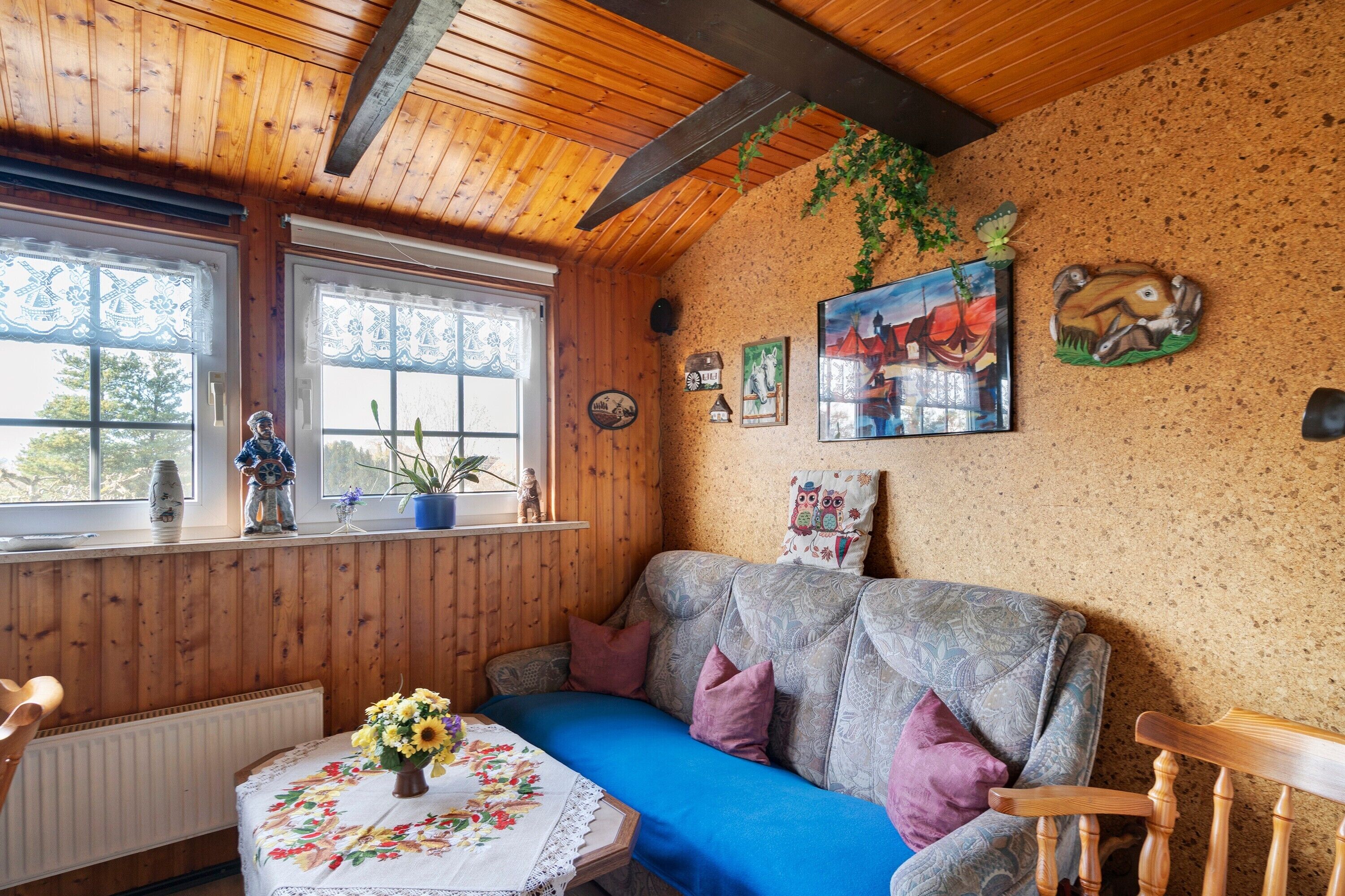 A cozy room with a wooden ceiling, a blue sofa, a small table with a floral tablecloth, and a window with lace curtains.