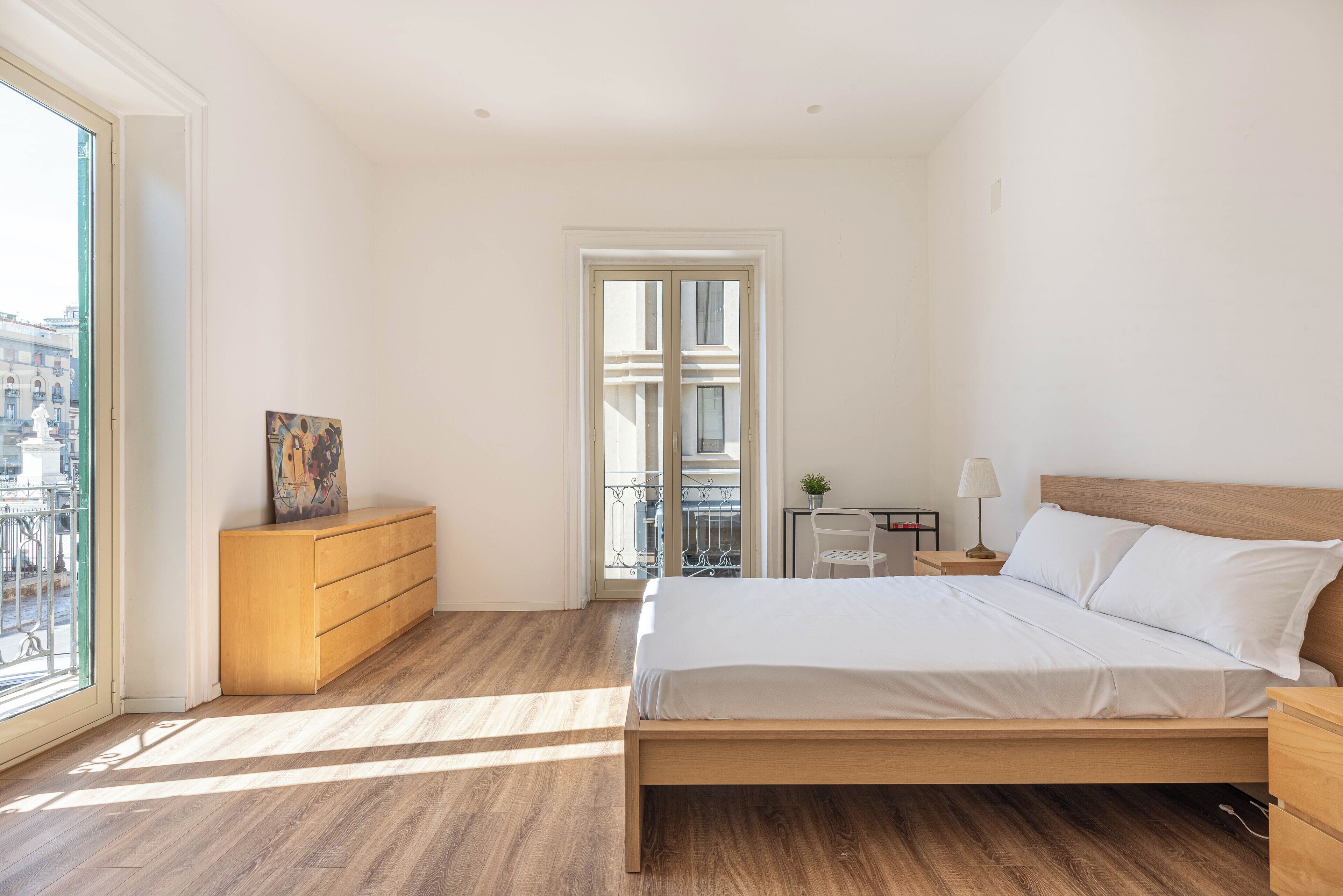 A bedroom with a wooden bed, a dresser, a nightstand, a lamp, and a view of the outside balcony.