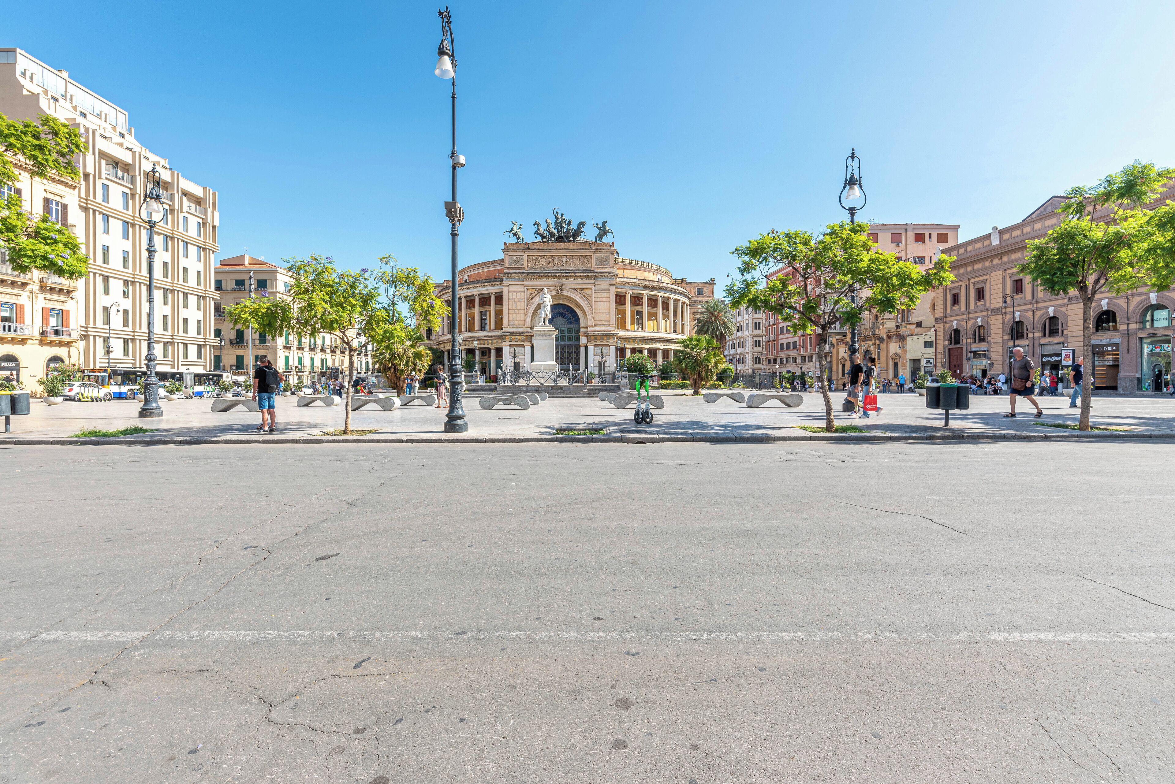 A wide urban square with a central historic building, surrounded by modern buildings and trees.