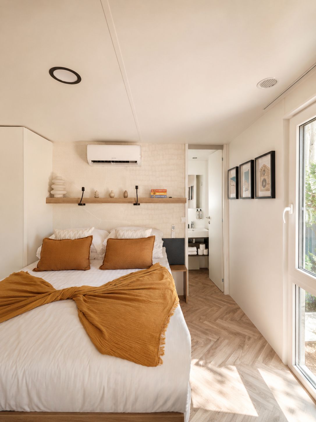 A modern bedroom with a large bed, a bedside table, a built-in shelf with decorative items, and a view of the bathroom through an open door.