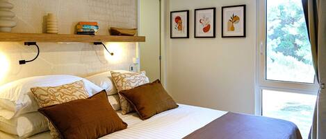 A neatly made bed with brown and patterned pillows, a wooden shelf with books and a decorative item, and two framed artworks on the wall.