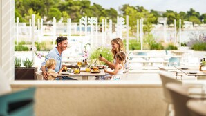 Une famille prend son repas dans un restaurant en plein air, avec vue sur un port de plaisance et sur la verdure environnante.