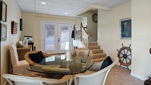 A dining area with wicker chairs and a glass table, a staircase, and a ship's wheel decoration.