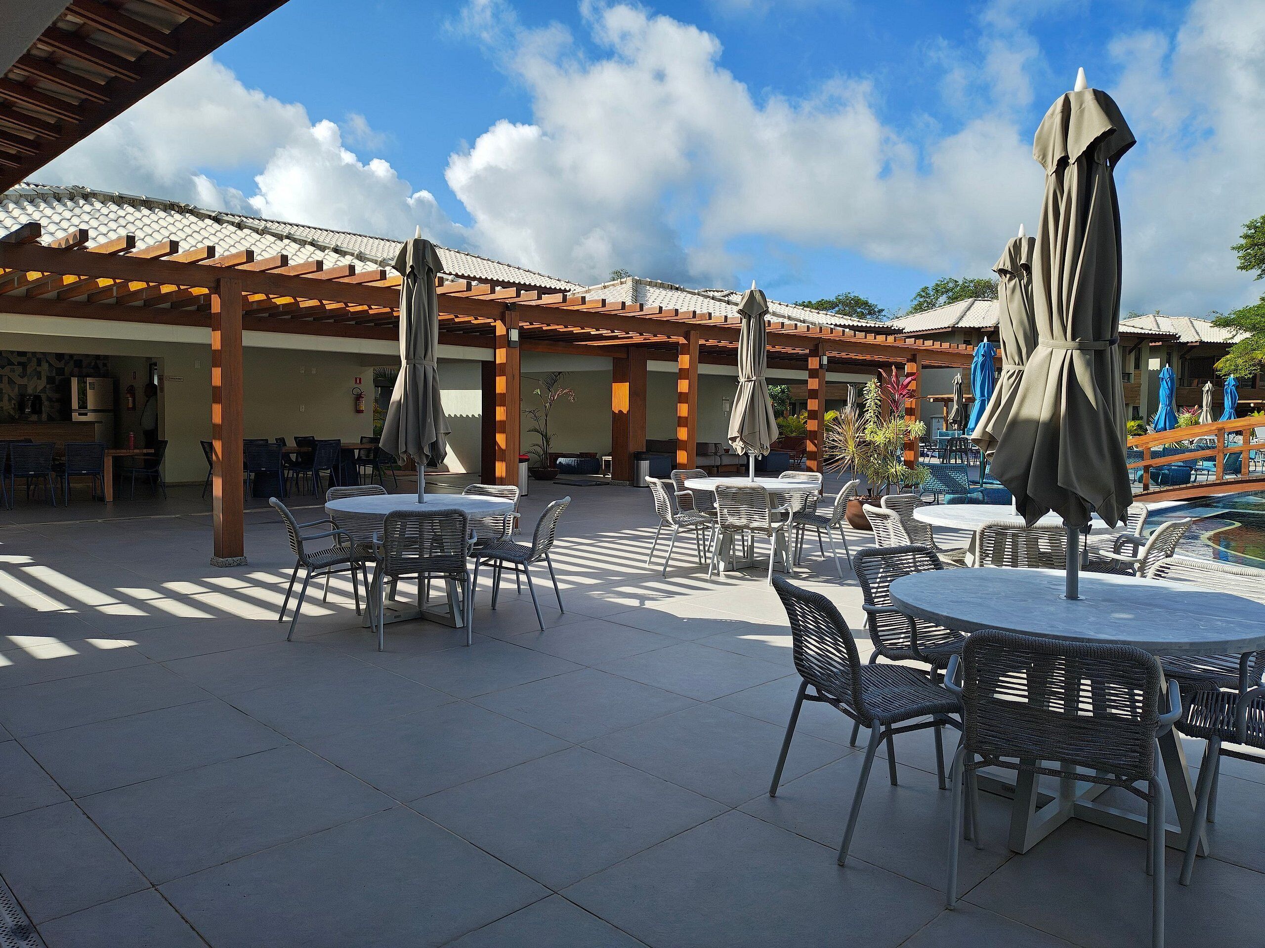 An outdoor patio area with round tables and chairs, covered by umbrellas, and a pool in the background.