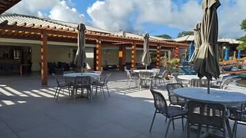 An outdoor patio area with round tables and chairs, covered by umbrellas, and a pool in the background.