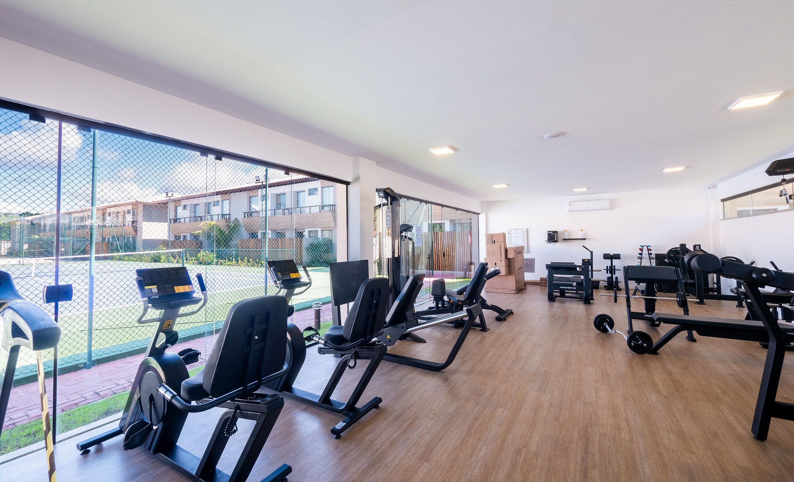 Uma academia com máquinas de cardio, equipamentos de musculação e uma grande janela com vista para um pátio.