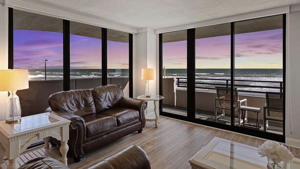 A modern living room with a leather sofa, a small table with a lamp, and a view of the ocean through large windows. - Horizons 202 (Daytona Beach)