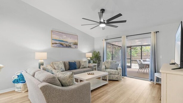 Ein modernes Wohnzimmer mit einer Couch, einem Couchtisch und einem Deckenventilator. An der Wand hängt ein Gemälde und ein Fenster ist mit Vorhängen versehen.
