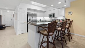 A modern kitchen with white cabinets, a stainless steel refrigerator, and a microwave. There are three wooden bar stools with cushioned seats. - Oceanwalk 11-603 (New Smyrna Beach)