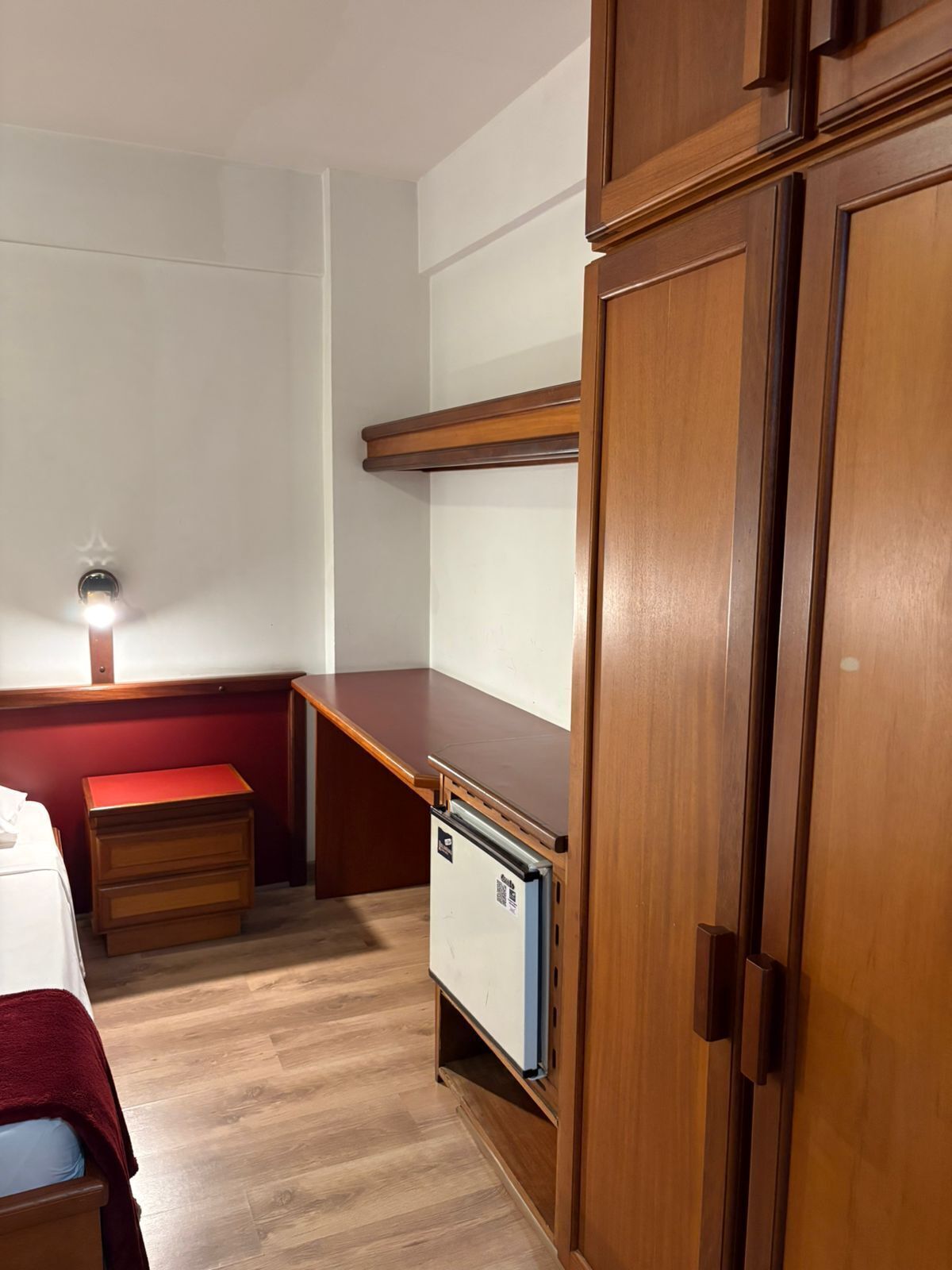 Ein kleines Zimmer mit einem Holztisch, einem kleinen Kühlschrank, einem Kleiderschrank und einem Bett mit roter Decke.