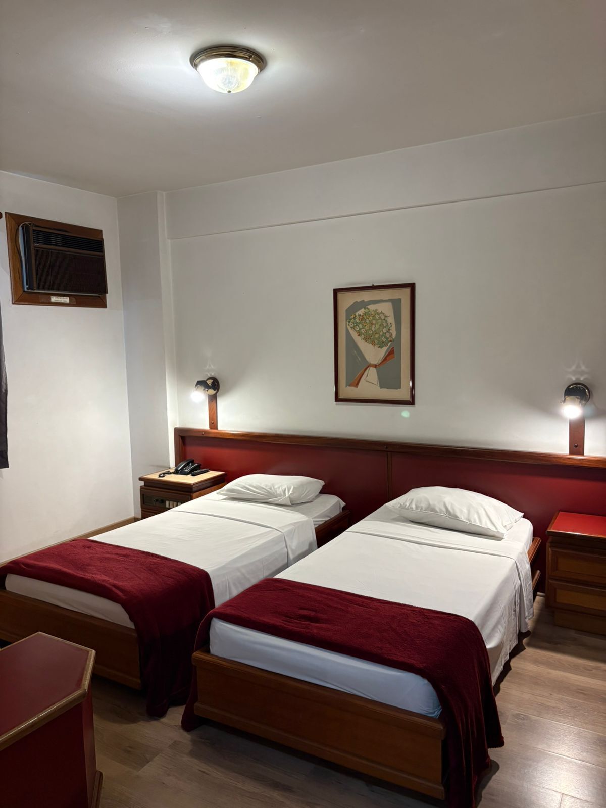 Una camera d'albergo con due letti singoli, una testiera in legno, un comodino con lampada, un telefono e un quadro appeso al muro.