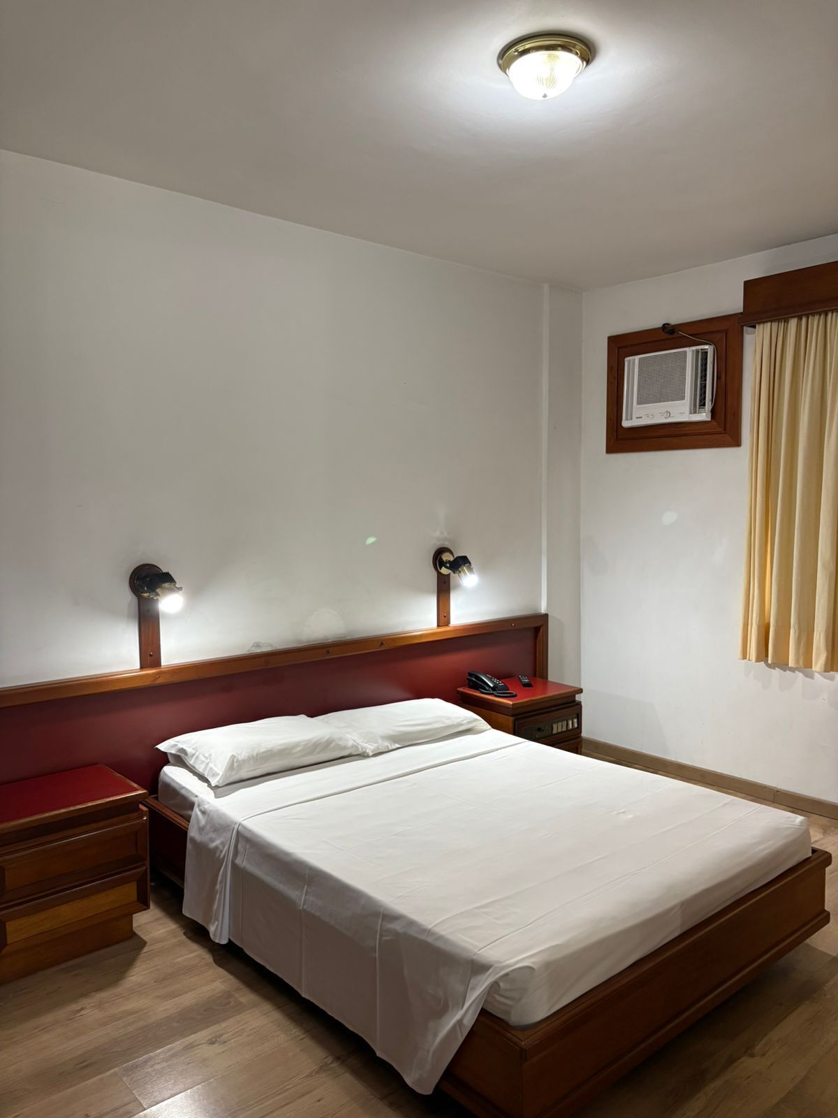 Een hotelkamer met een eenpersoonsbed, houten hoofdeinde, nachtkastjes, een wandairconditioner en een raam met gordijnen.