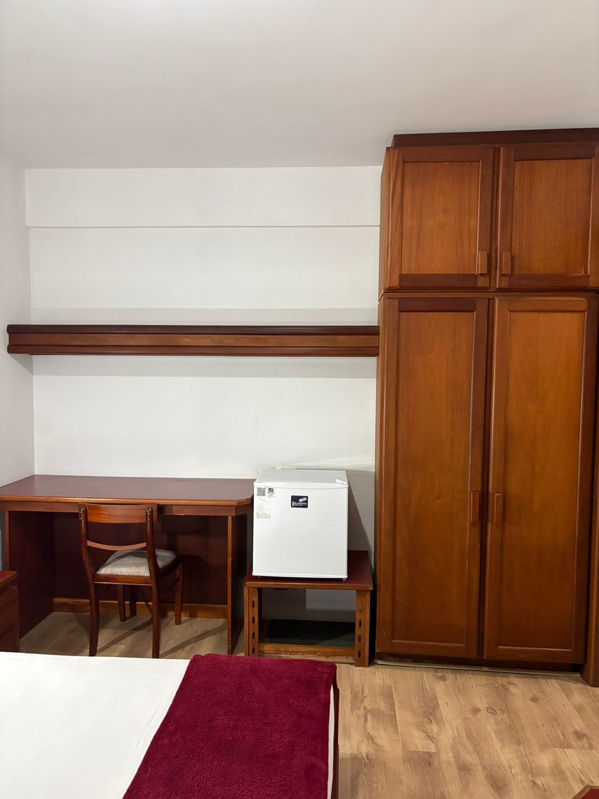 A room with a wooden desk, a chair, a small white refrigerator, and a wardrobe.