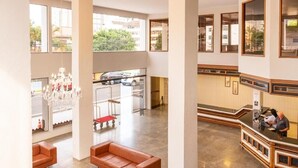 A spacious lobby with high ceilings, a chandelier, leather sofas, a reception desk, and large windows offering views of the street. - Goldmen Business Hotel Blumenau (Blumenau)