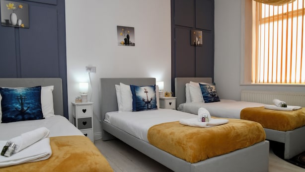 A hotel room with two beds, each with a white headboard and a blue pillow. There are two bedside tables with lamps, a window with blinds, and a small painting on the wall.