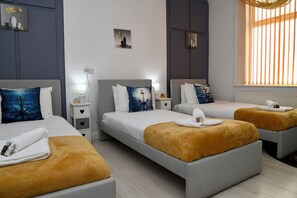 Une chambre d’hôtel avec deux lits, chacun doté d’une tête de lit blanche et d’un oreiller bleu. On y trouve deux tables de chevet avec des lampes, une fenêtre avec des stores et un petit tableau accroché au mur.