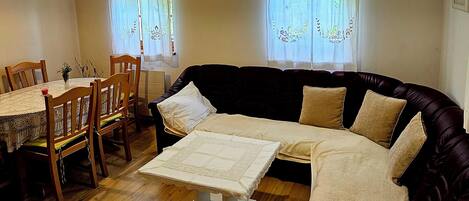 Ein Wohnzimmer mit einem dunkelgrauen Sofa, einem kleinen Tisch mit weißem Tuch, Holzstühlen und einem Esstisch.