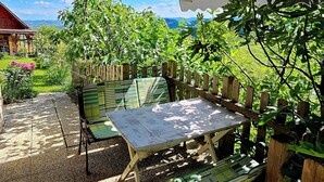 En udendørs patio med bord og stole under et skyggefuldt halvtag, omgivet af grønt og med udsigt til bjergene.