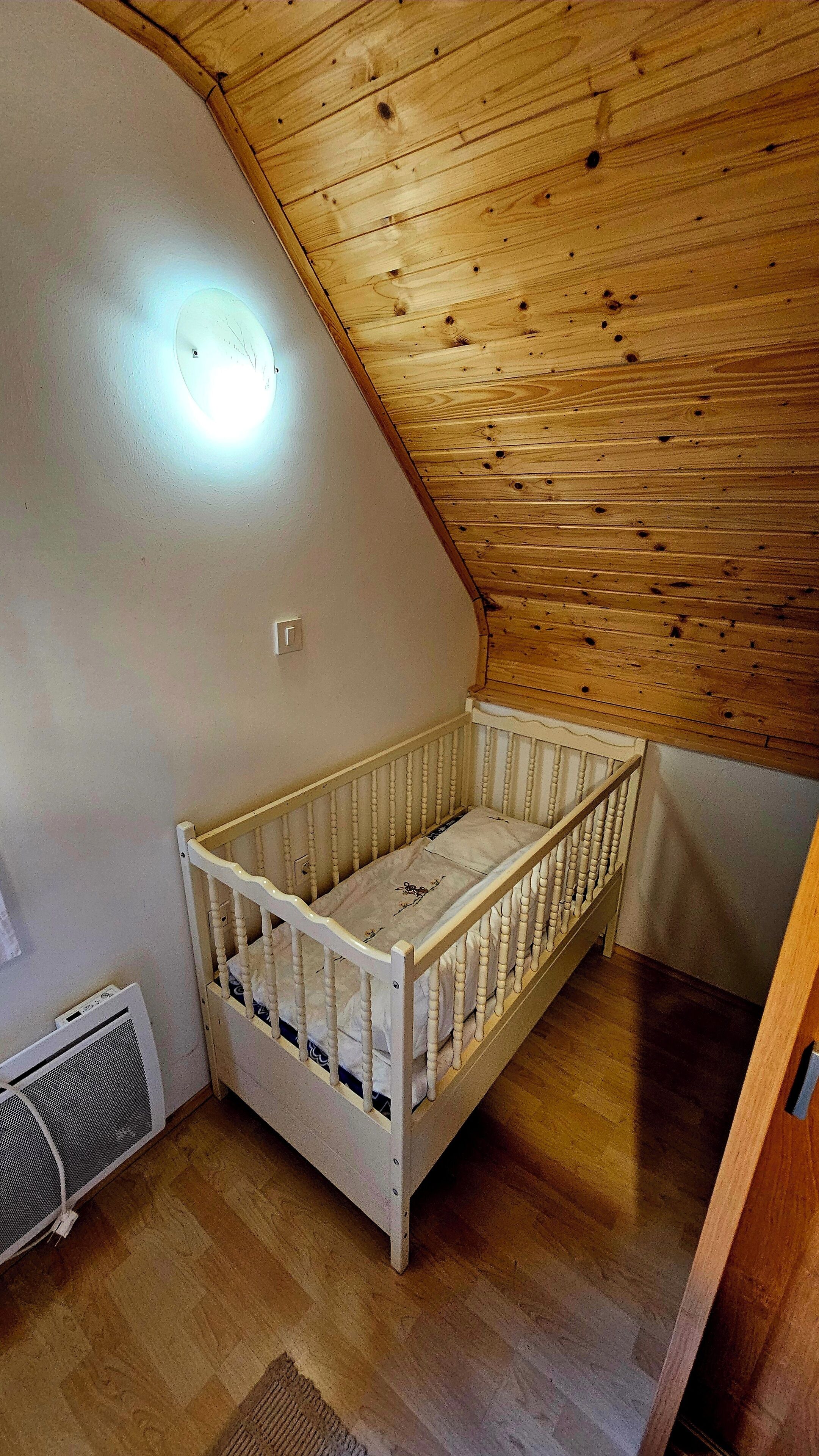 A small, single-room apartment with a sloped ceiling, a white crib, a heater, and a radiator on the floor.