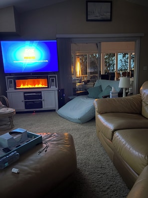 A living room with a flat-screen TV, a fireplace, a sofa, and a dining area visible through a glass door. - Beautiful lake house with stunning views of  Pere Marquette and Lake Michigan (Ludington)