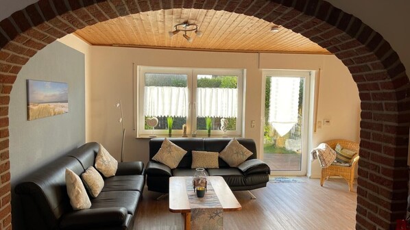 Un salon avec un canapé en cuir noir, une table basse en bois et un fauteuil en osier. Il y a deux fenêtres avec des rideaux, une arche en briques et un ventilateur de plafond.