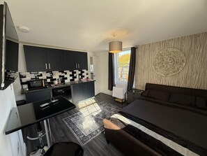 A compact hotel room with a kitchenette, a bed, a large window with curtains, and a decorative wall piece. - Aspendos Hotel (Sturry)