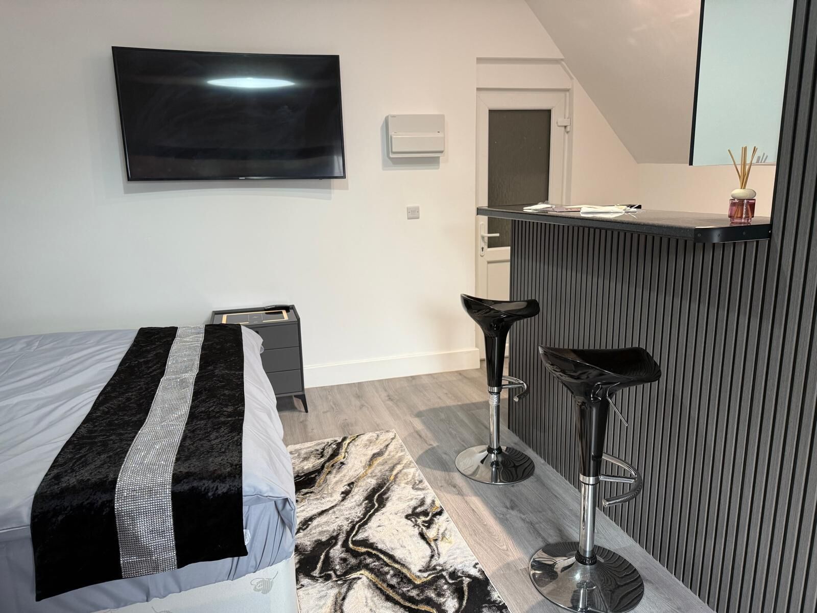 A modern hotel room with a bed, a TV, a minibar, and two bar stools.