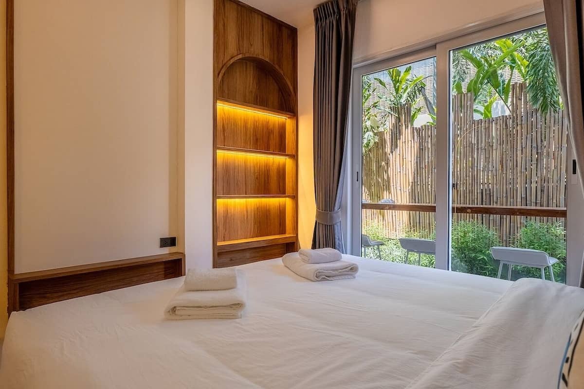 A bedroom with a bed, a wooden headboard with built-in lighting, a window with a view of greenery, and a small table by the window.