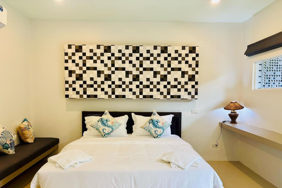 A neatly made bed with white linens and decorative pillows, a checkered wall art above, and a bedside lamp on a nightstand.