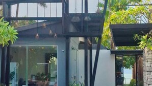 A modern building with a glass facade, a covered outdoor seating area, and a stone wall. - The Strand Resort Samui  (Koh Samui)